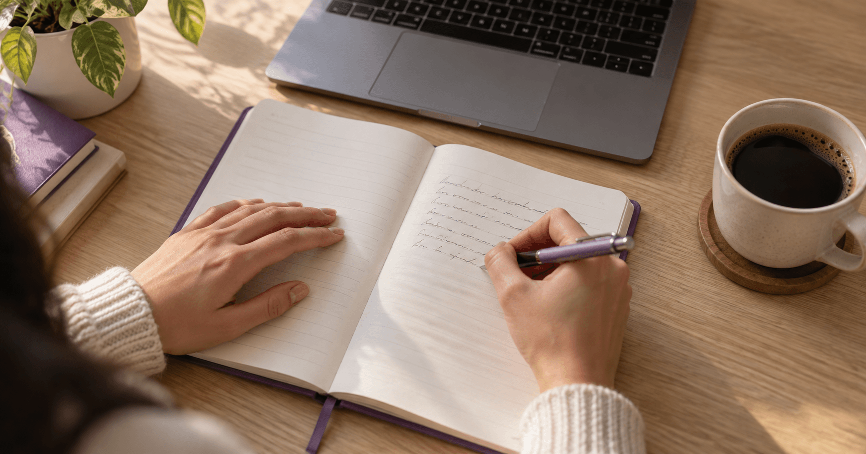 Manos escribiendo una carta en una libreta junto a un computador portátil y una taza de café